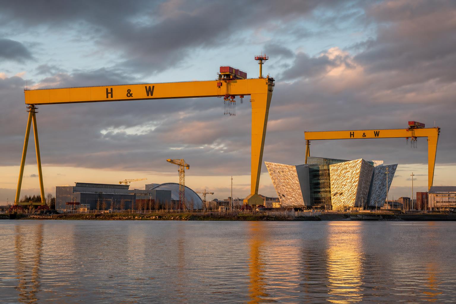 Belfast skyline at golden hour — the yellow Harland & Wolff Samson and Goliath shipyard cranes, the angular Titanic Belfast building reflecting warm light over the calm River Lagan